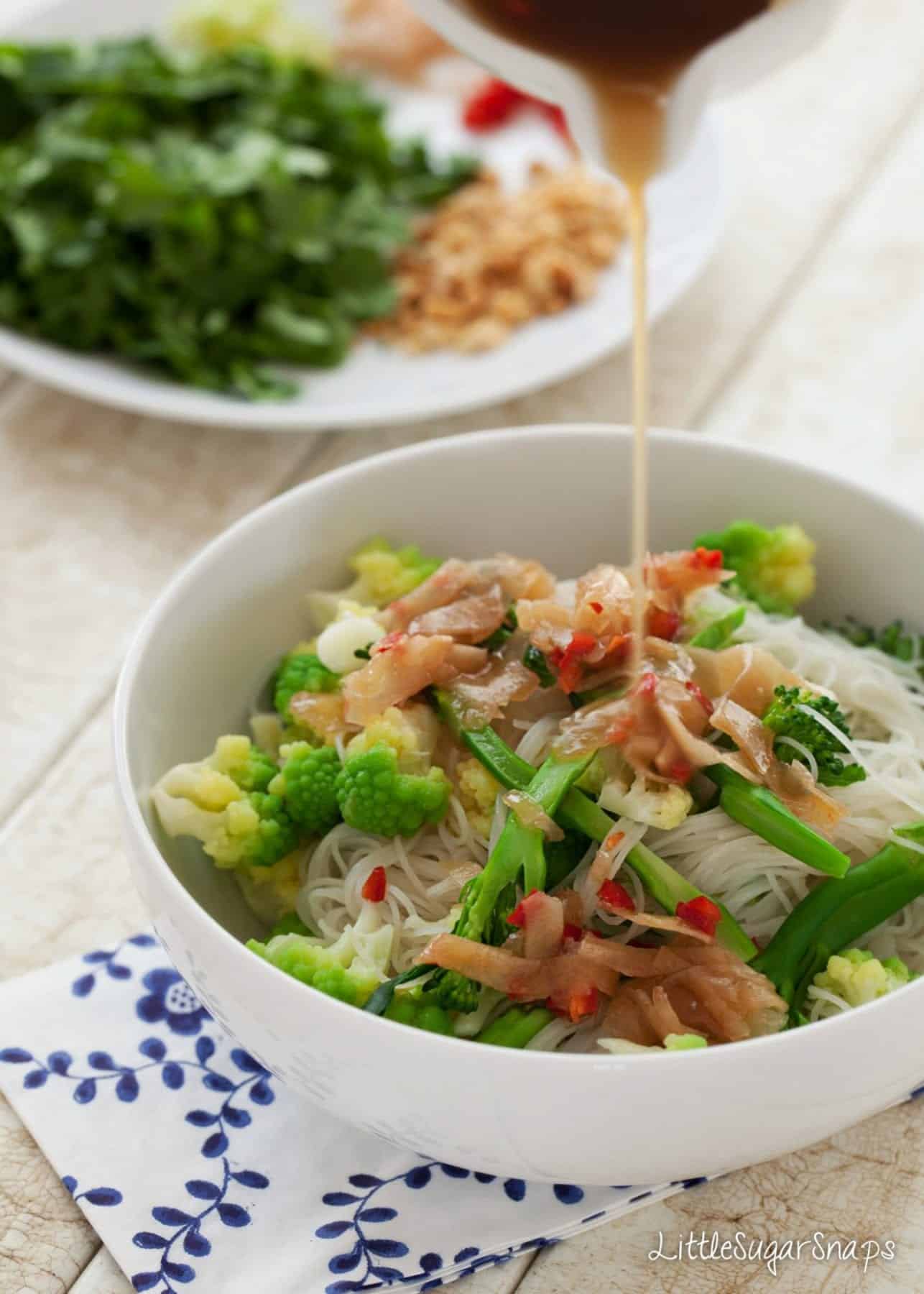 Dressing being poured onto a bowl of noodle salad with pickled ginger, and broccoli