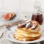A stack of ricotta & almond pancakes serves with figs, almonds and syrup and duated with icing sugar. Served on a blue & white plat which is sitting on a black & white comic strip