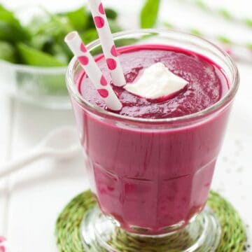 A glass of beetrootsmoothie with paper straws in it, a dollop of yoghurt and some chai seeds on the worktop