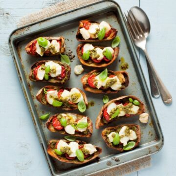 Ten loaded Potato Skins with Pesto and Goats Cheese on a baking sheet which is on a piece of cloth with serving utensils next to it.