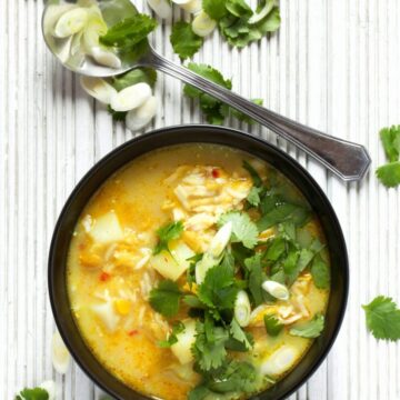 Split Pea, Chicken & Rice soup in a black bowl. Lots of fresh coriander and spring onion slices has been scattered on top and these ingredients are also on the wooden tabletop the bowl is on