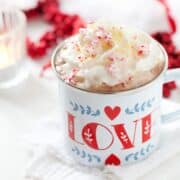 An enamel mug (with the word love on it) of hot chocolate with cream, crushed candy cane pieces and popping candy. Christmas decorations in the background