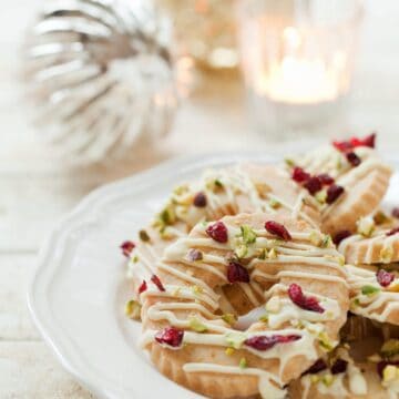 Christmas wreath biscuits topped with white chocolate, cranberries and pistachio nuts. Served on a white plate with baubles in the background