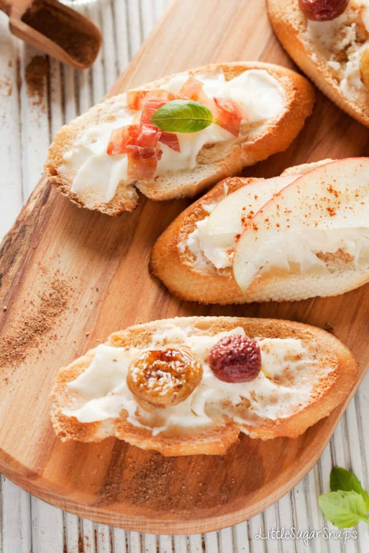 Crostini on a wooden board topped with taleggio, apple, grapes and ham