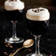A glass of coffee, blackcurrant and almond liqueur drink topped with softly whipped cream. On a black tablecloth with coffee beans scattered around