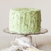 A cake covered in green icng with chocolate pieces in it to give the look of mint choc chip. They cake is on a glass stand with a cloth wrapped around it