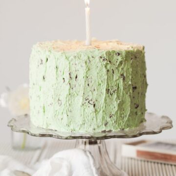 A chocolate cake covered in green mint buttercream with chocolate pieces in it. The cake has a single lit candle in the centre