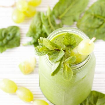 A green smoothie topped with grapes and mint and ingredients surrounding it on the white wooden worktop