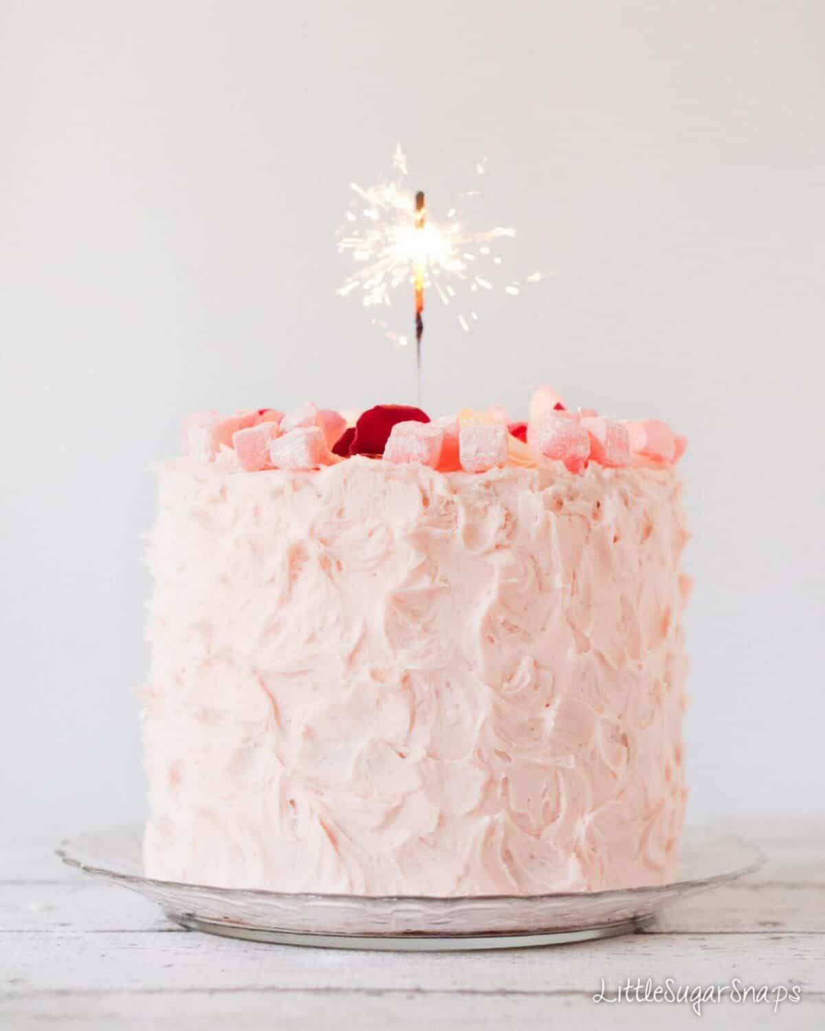 A celebration cake covered in pink frosting and decoated with turkish delight chunks and rose petals with a lit sparkler in the centre