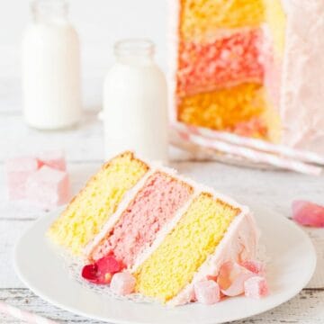 A lemon and rose triple layer cake cut open with a slice on a plate in focus at the front of the shot. Two yellow lemon layers and one pink rose layer are sandwiched together with pink icng and decorated with turkish delight and rose petals