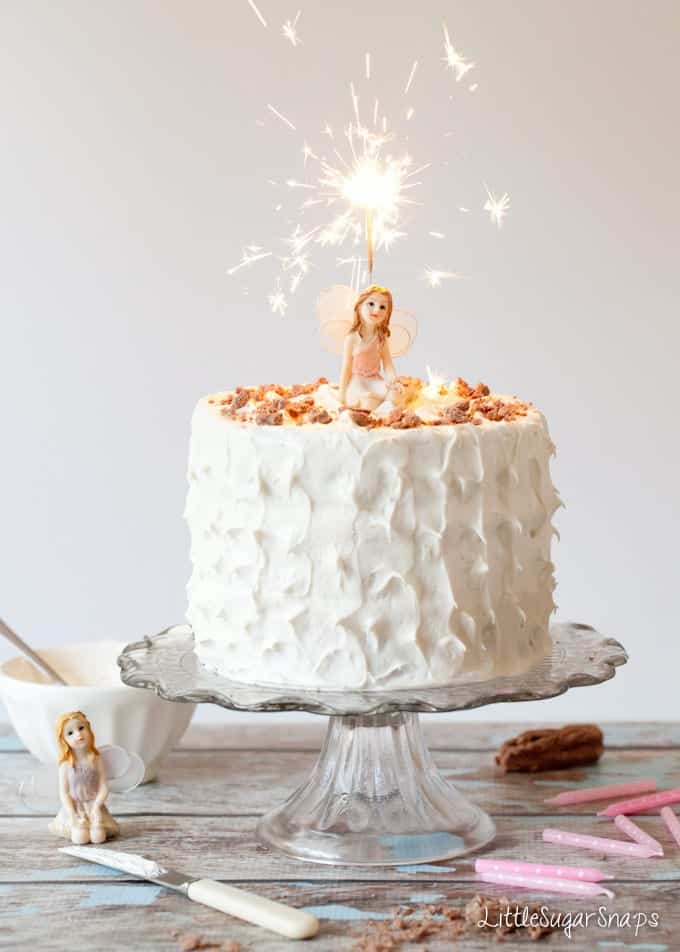 Three layer cake with marshmallow frosting, a lit sparkler and a fairy ornament.