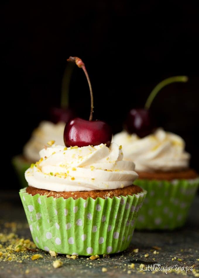 Pistachio Cupcakes in green wrapper with frosting, chopped pistachios and fresh cherries.