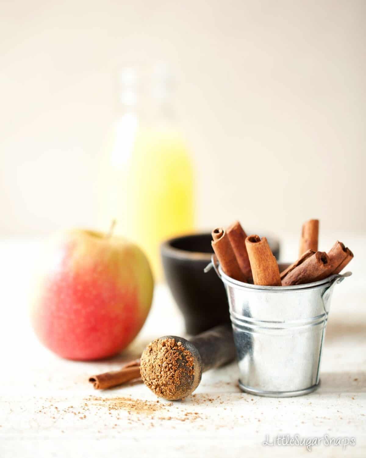 A small bucket of cinnamon sticks with ground cinnamon on the end of a pestle, an apple and fruit juice