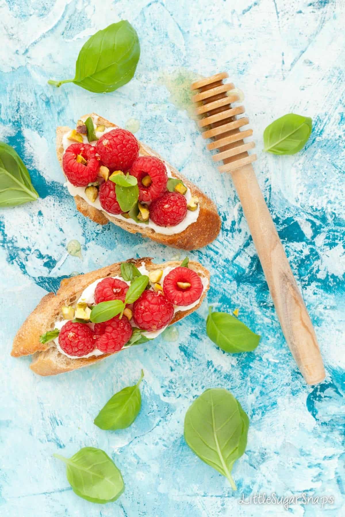 Overhead of fruit breakfast bruschetta: toasted baguette topped with soft cheese, raspberries, pistachio and basil