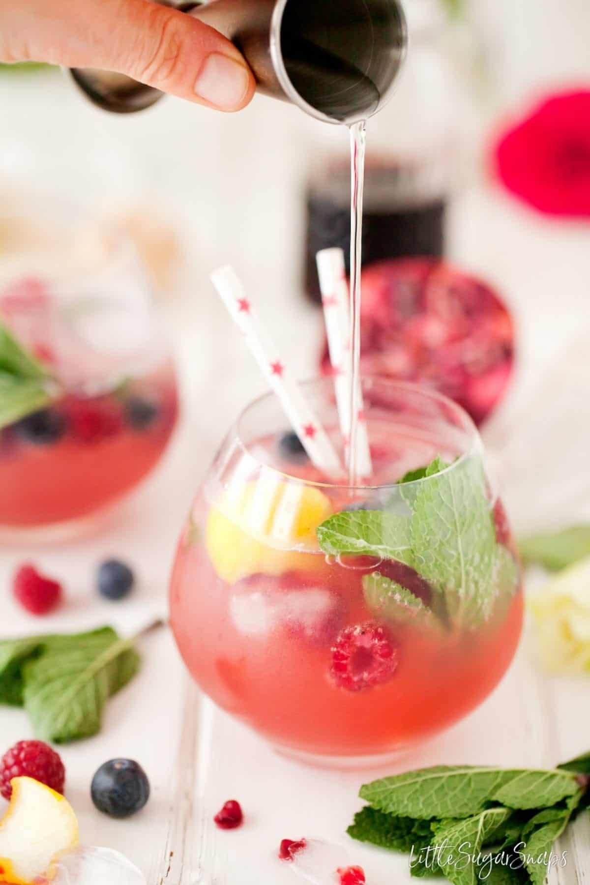 Person pouring a measure of vodka into a fruity grenadine drink.