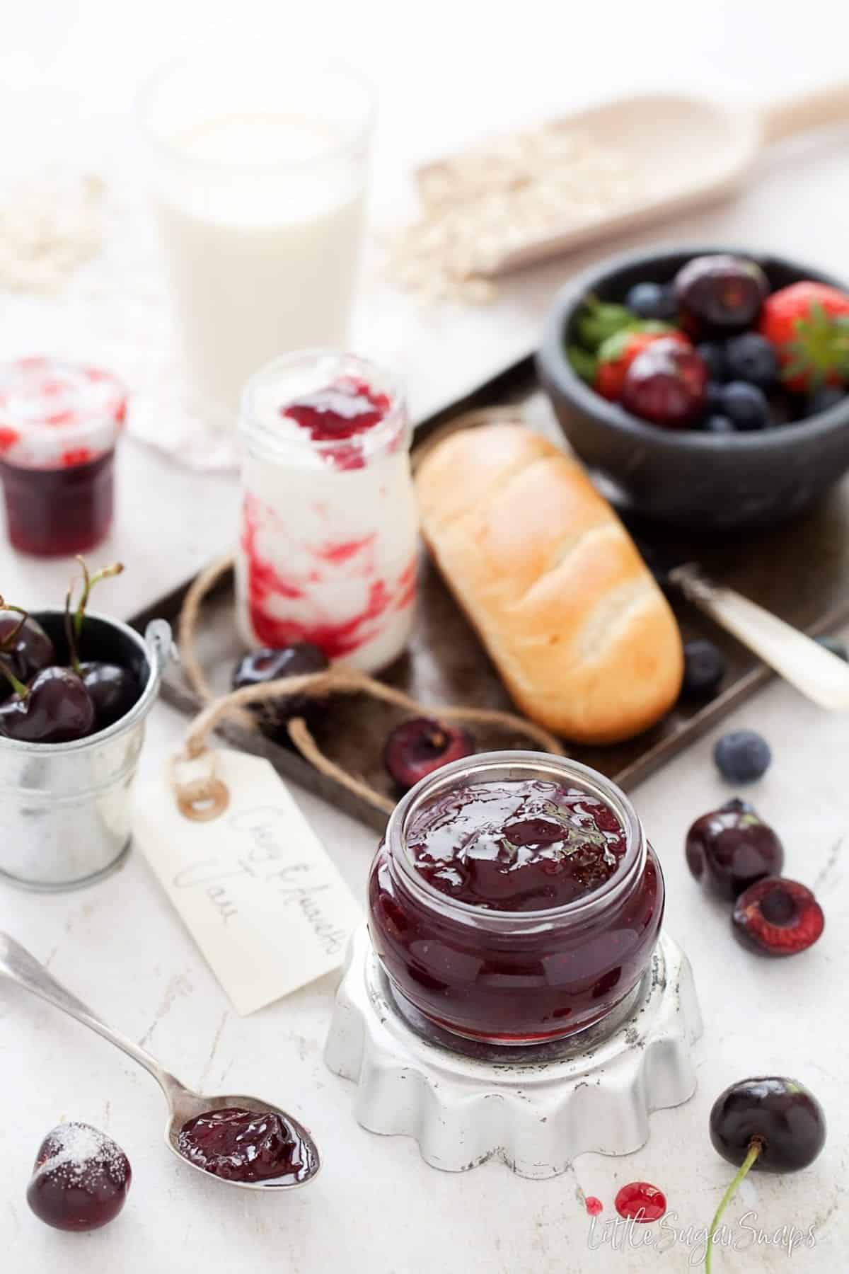 A jar of Amaretto Cherry Jam with fresh cherries and brioche rolls