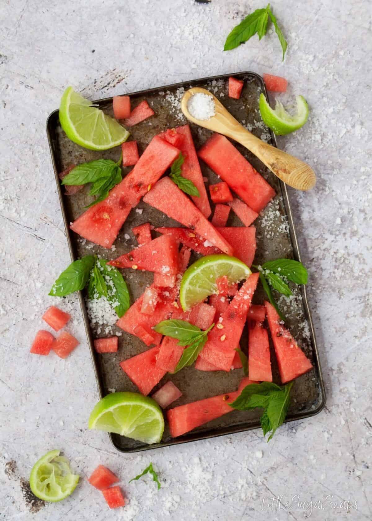 Wedges of watermelon on a baking tin with mint, lime and salt.