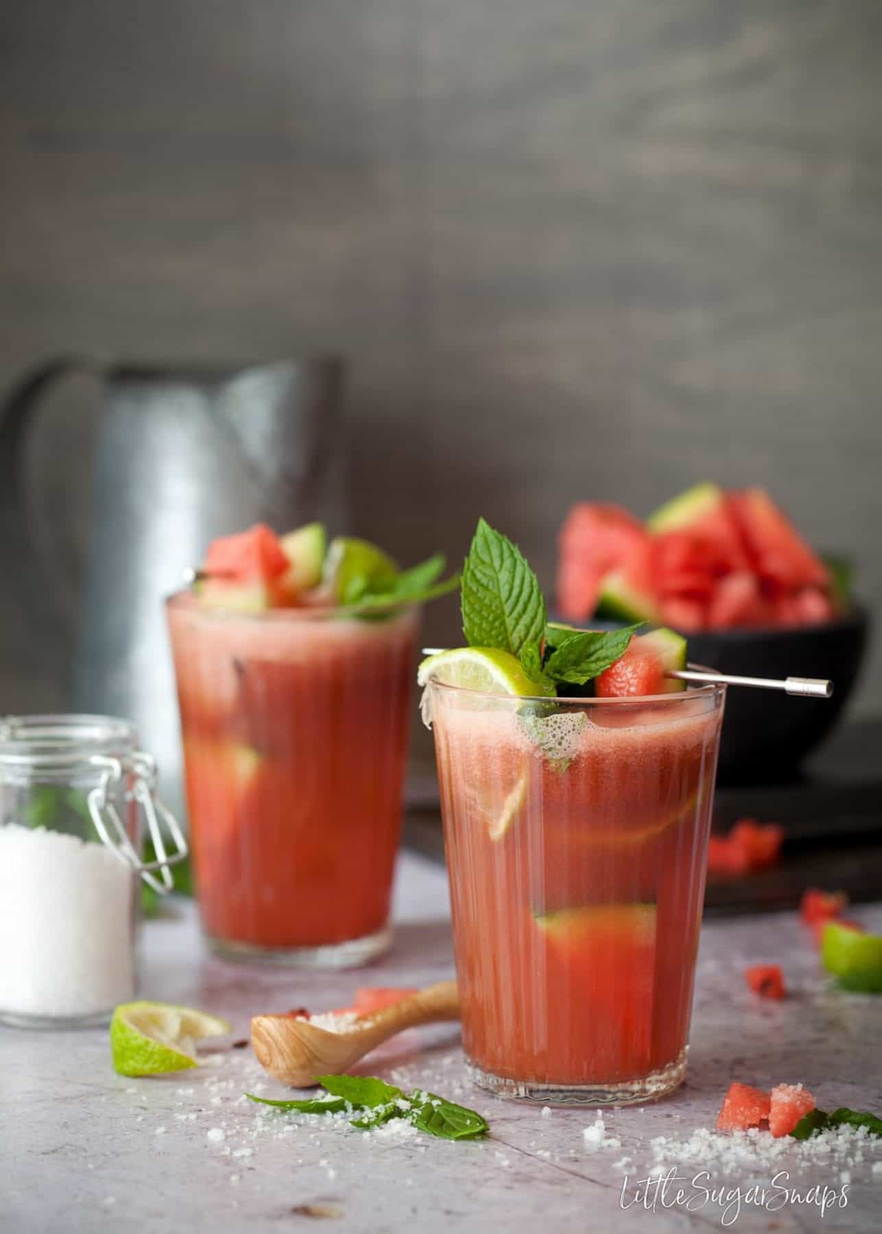 Two glasses of watermelon juice garnished with lime, mint and fruit.