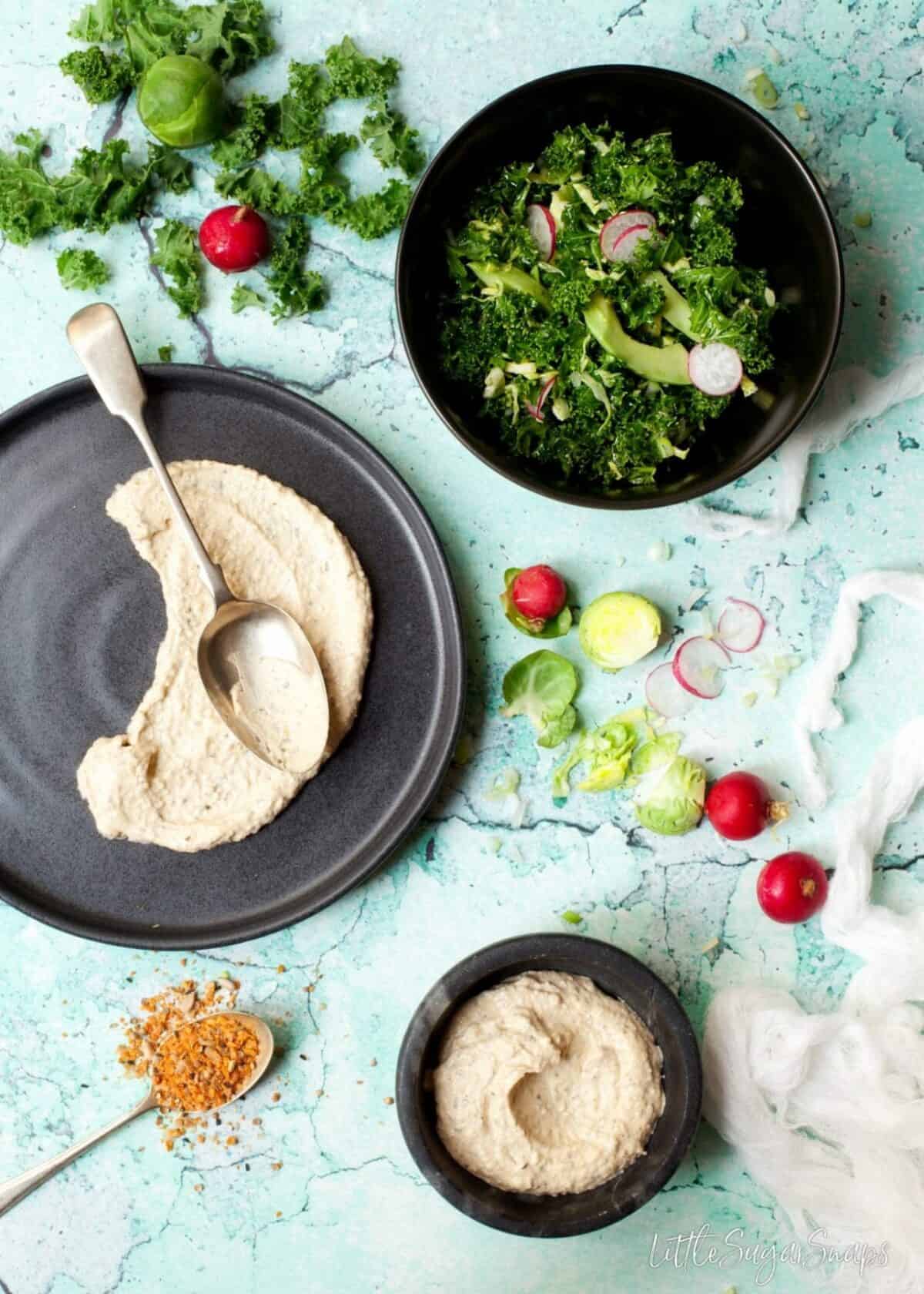 Hummus spread onto a plate with salad ingredients in shot.