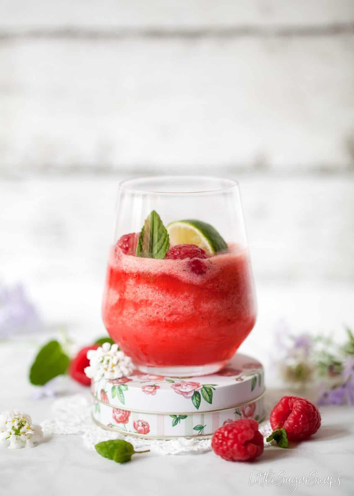 A glass of frozen gin & tonic with raspberries and mint on a floral tin