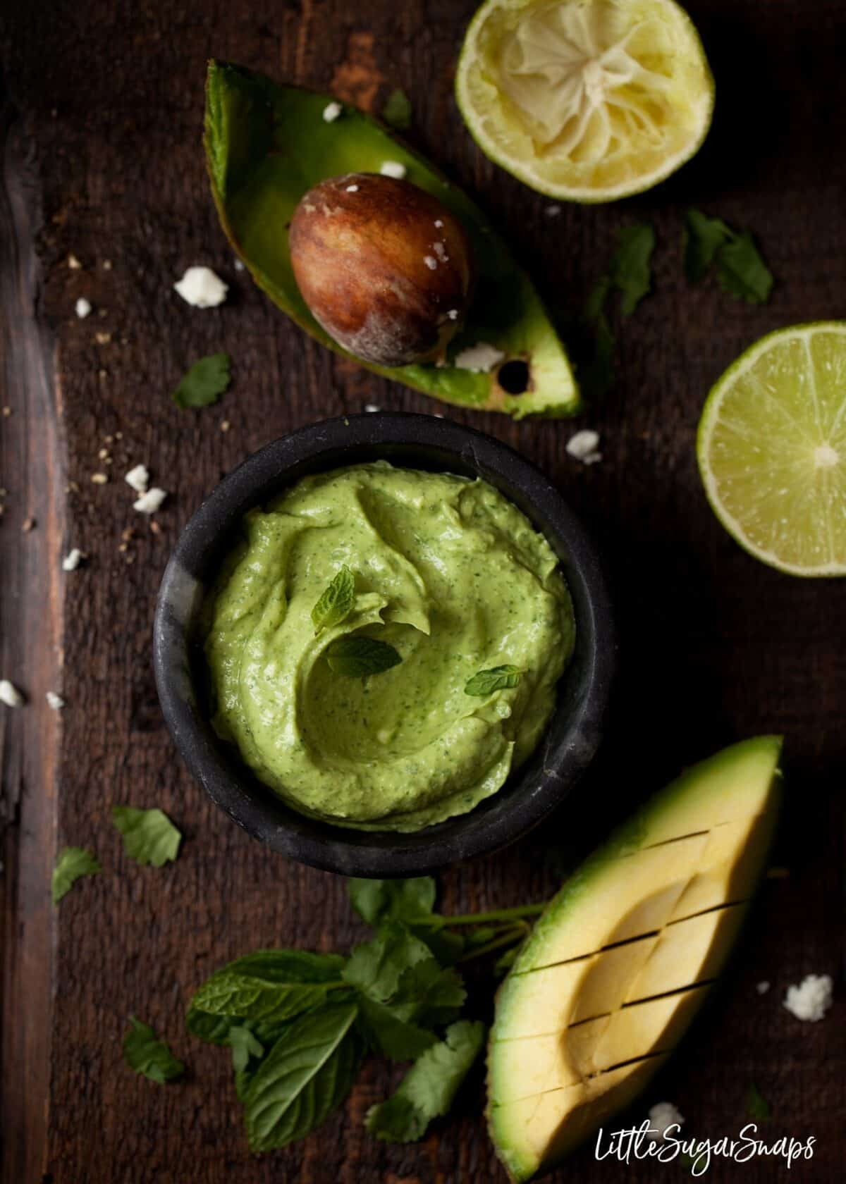 A bowl of avocado and feta dip with ingredients alongside.