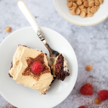 Cutting into raspberry brownie with peanut butter frosting. The brownie is in a bowl with a fork resting inside. On the worktop is a small bowl of peanut butter candy chips and fresh raspberries scattered around