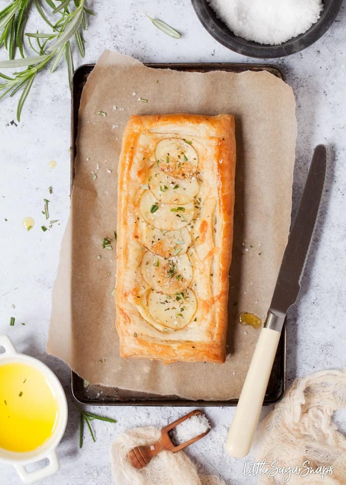 a cheesy potato tart on a baking sheet with rosemary, sea salt, olive oil and a knife for slicing