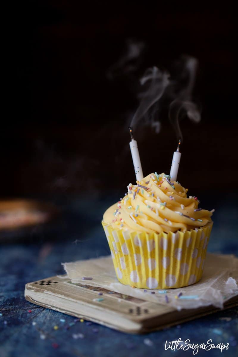 A cupcake in a yellow wrapper topped with french buttercream, sprinkles and 2 smoking candles. 