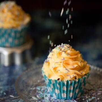 Cupcakes topped with piped French buttercream with sprinkles being scattered on top. The cake in in a bright blue wrapper with polka dots