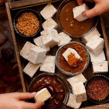 A tray filled with pumpkin spice marshmallow chunks, chocolate sauce, caramel sauce, crushed cookies, bronze sprinkles and caramel curls. Children are dipping some of the marshmallow into the sauces