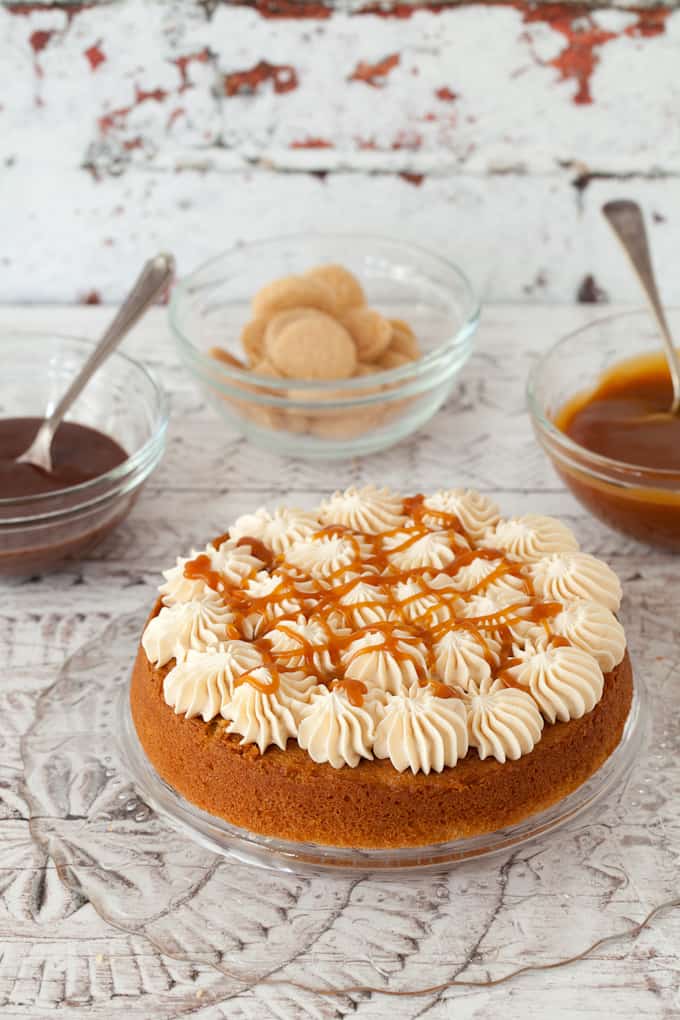 decorating a millionaire's cake. bottom layer of sponge cake has rosettes of buttercream on top and caramel sauce drizzled over it. Pots of caralem & chocolate sauce behind along with shortbread cookies