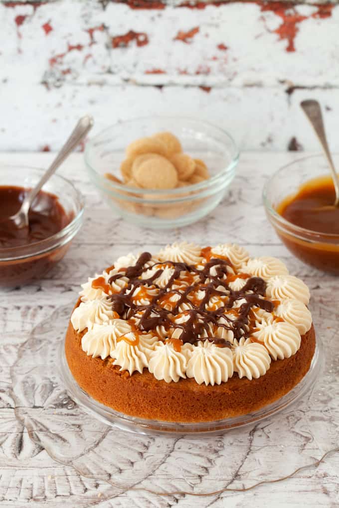 decorating a millionaire's cake. bottom layer of sponge cake has rosettes of buttercream on top and caramel & chocolate sauce drizzled over it. Pots of caramel & chocolate sauce behind along with shortbread cookies