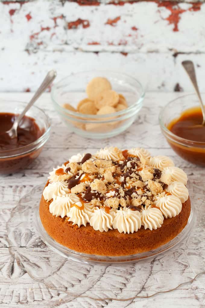 decorating a millionaire's cake. bottom layer of sponge cake has rosettes of buttercream on top, chocolate & caramel sauce and chunks of shortbread cookies on top. Pots of caramel & chocolate sauce behind along with shortbread cookies