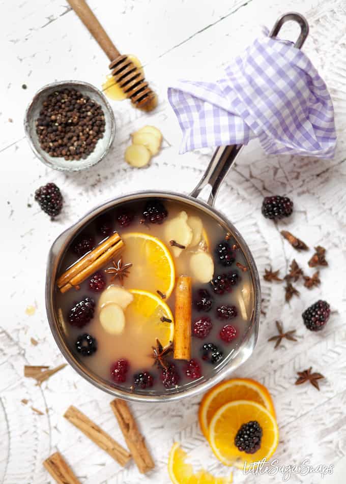 A pan filled with apple juice and mulling ingredients like cinnamon and star anise, orange slices and blackberires still in the pan and ingredients scattered around