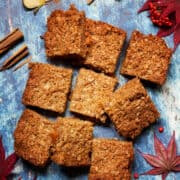 Nine pieces of flapjack on a blue background with slices of apple, cinnamon and autumn leaves and berries surrounding it