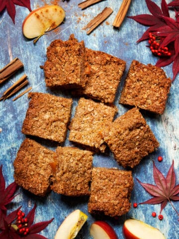 Nine pieces of flapjack on a blue background with slices of apple, cinnamon and autumn leaves and berries surrounding it