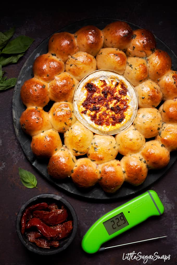 Bread rolls on a serving plate surrounding baked cheese that has been topped with red pesto.

