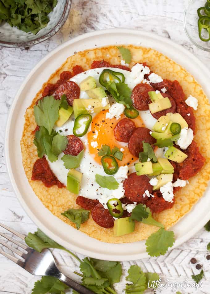 Overhead of cornmeal pancake with a fried egg, salsa, avocado, green chilli, feta and chorizo.