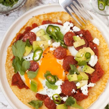 Huevos Rancheros with a cornmeal Crepe with spicy tomato sauce, a fried egg, green chillis and coriander plus chorizo, feta cheese and avocado chunks. The egg yolk has been broken open.