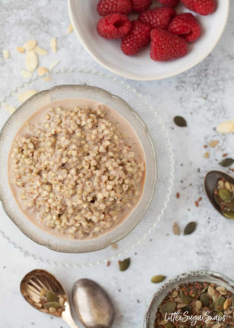 A bowl of spiced buckwheat porridge with raspberries, seeds and nuts alongside.