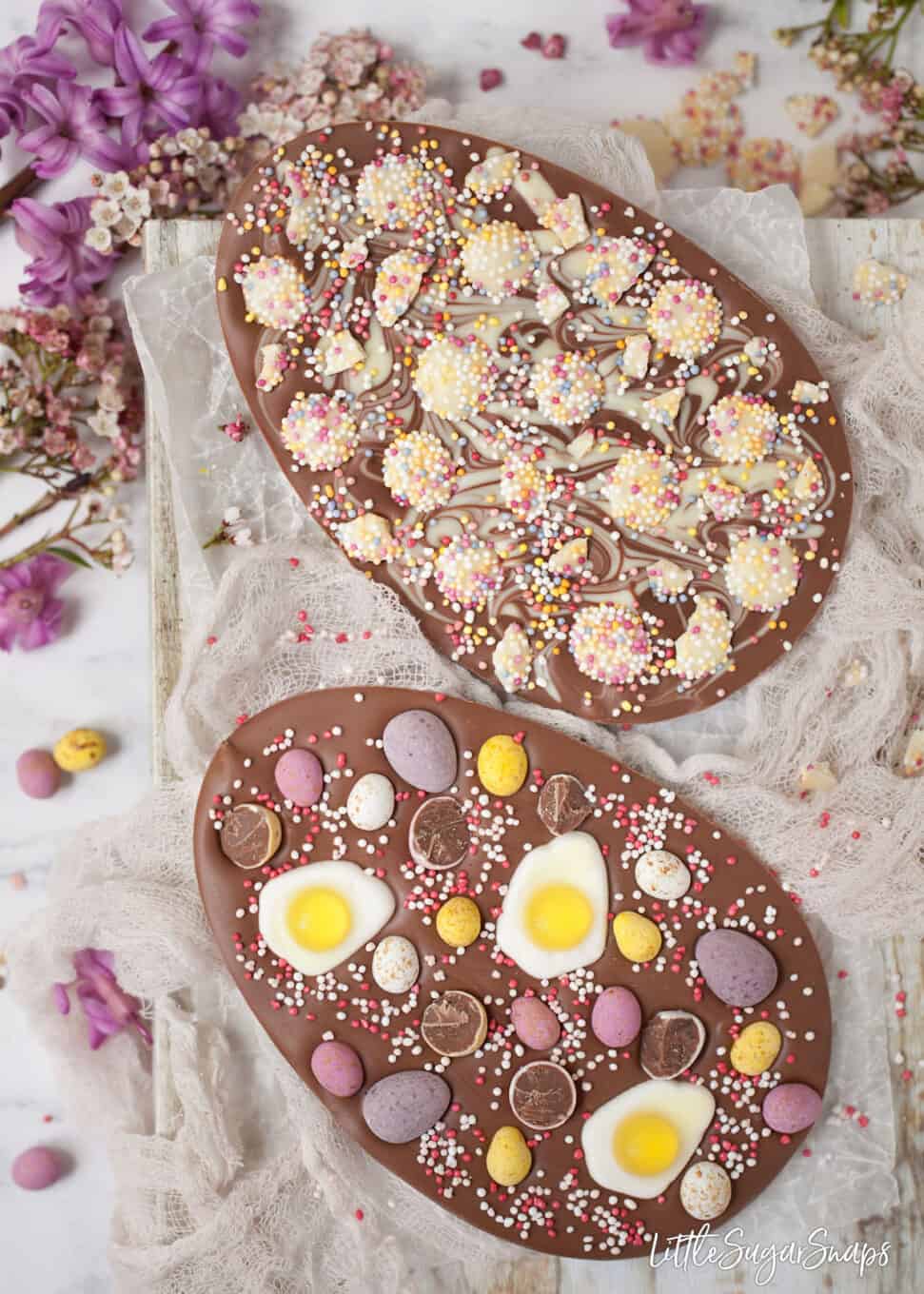 Chocolate eggs topped with mini eggs, sprinkles and jelly sweets. On cloth, with flowers in the background
