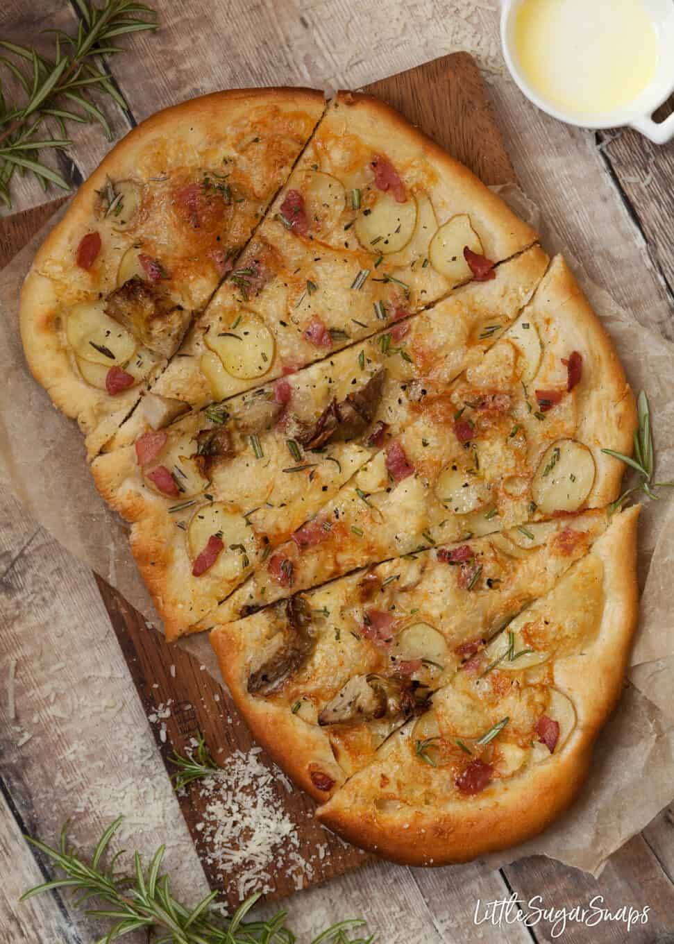 Cooked pizza topped with potato, cheese, bacon and rosemary on greaseproof paper with a wooden board underneath