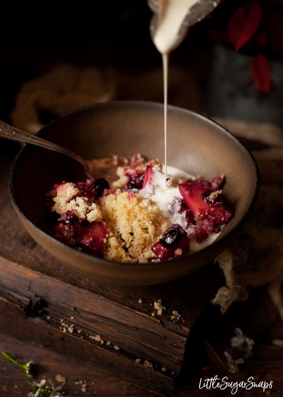 Cream being poured from a glass jug over an apple and blackcurrant crumble.