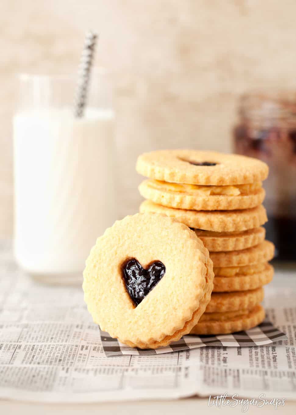 Biscuits made from a custard creams recipe and filled with custard cream filling and jam. Stacked up next to a glass of milk with one on it's side in front of the stack