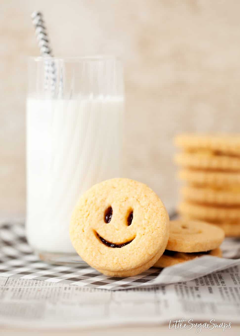 Homemade Jammie dodger with a smiley face front next to a glass of milk. More cookies in the background