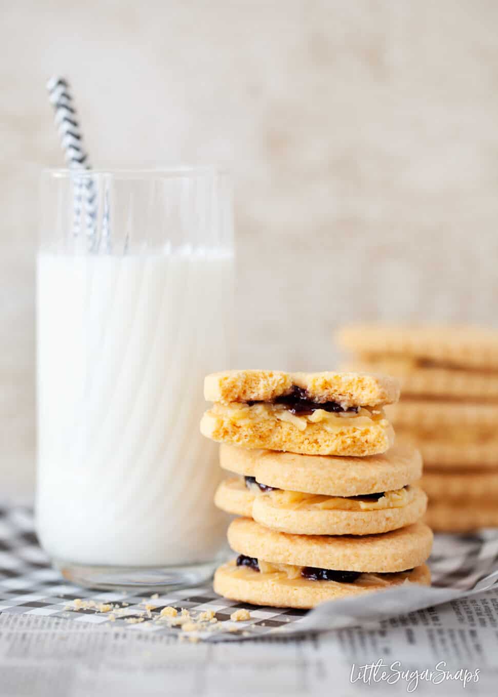 Biscuits made from a custard creams recipe stacked next to a tall glass of milk. Top cookie is bitten into to reveal the cream filling and jam