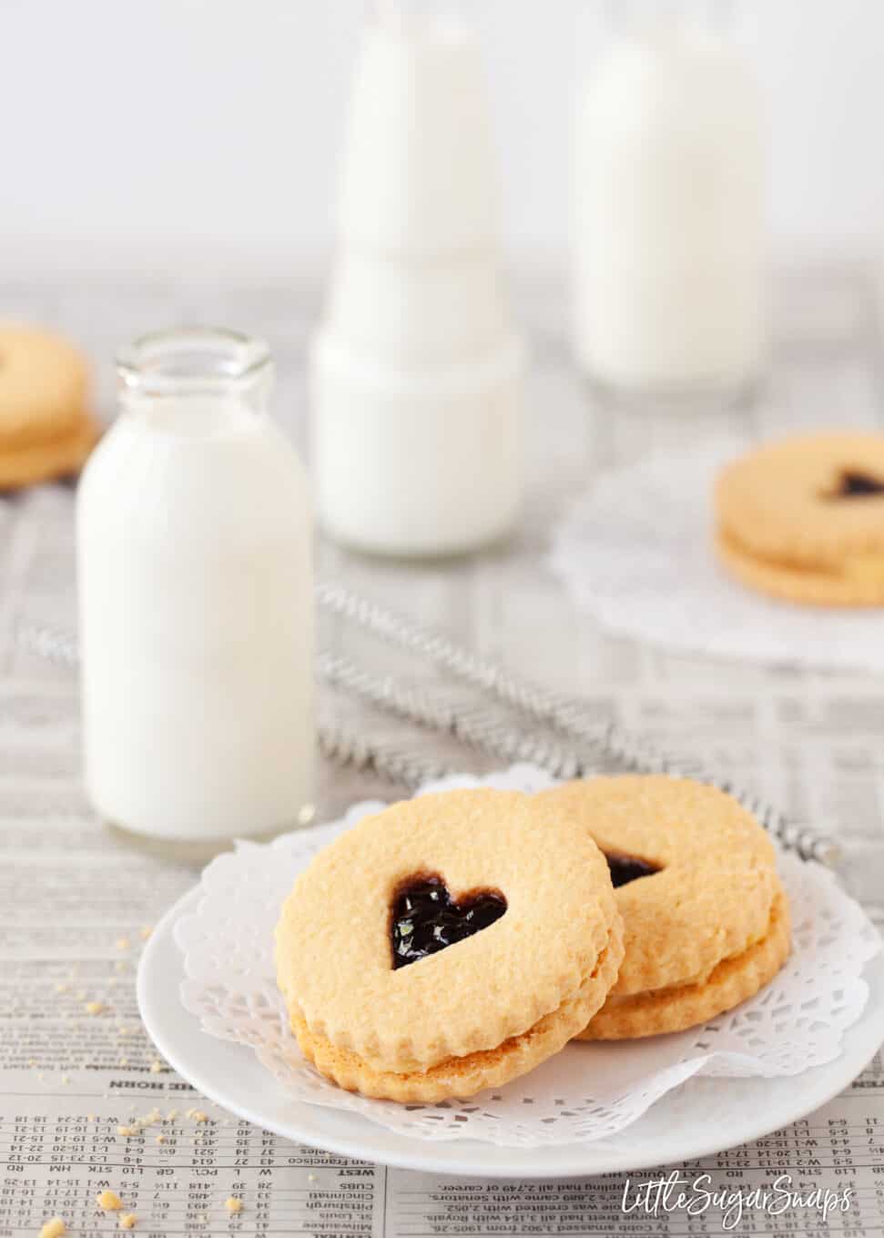 homemade Jammie Dodger cookies on a small plate with mini bottles of milk behind - with a love heart centre cut out to reveal the jam