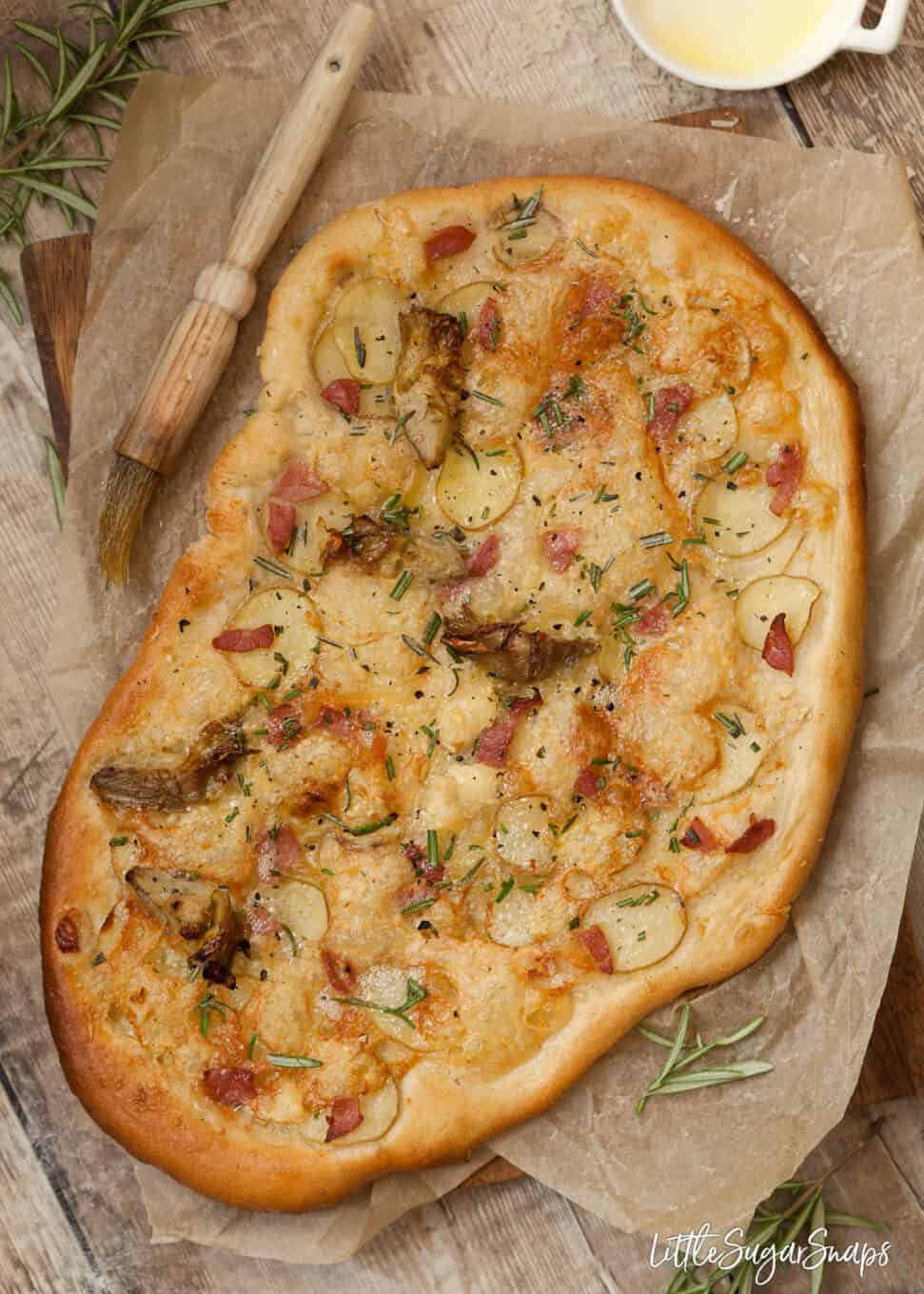 Pizza bianca without tomato sauce topped with sliced potato, bacon, artichoke, cheese and rosemary. Pastry brush alongside which has just been used to wipe olive oil around the edge of the pizza