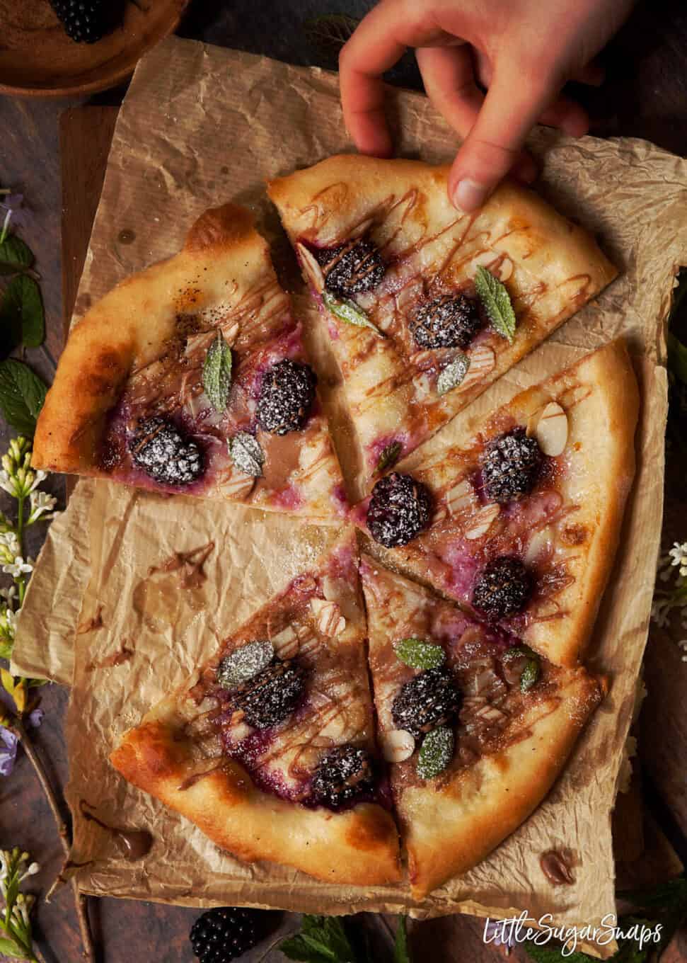 Person taking a slice of sweet dessert pizza topped with blackberries, chocolate drizzle and fresh mint.
