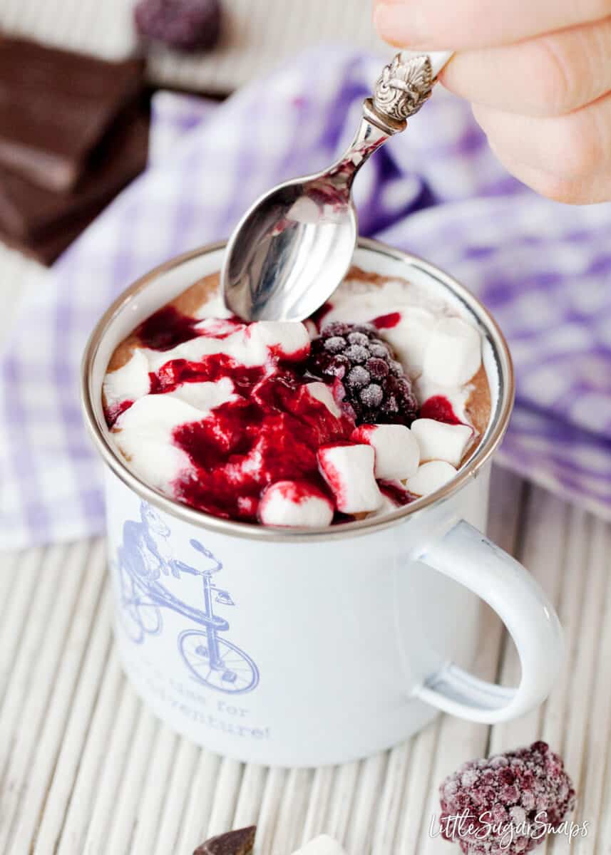 Person about to dip a spoon into hot chocolate with cream, blackberry coulis and marshmallows.